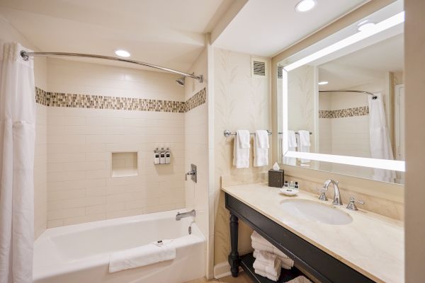 A modern bathroom with a tub, shower curtain, towels, a sink on a marble countertop, and a large mirror with built-in lighting.