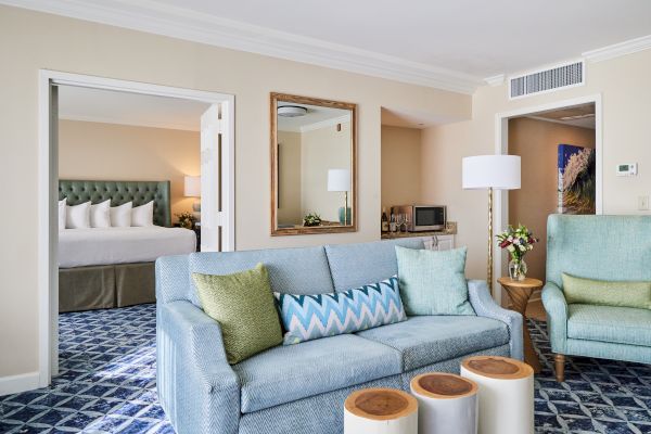 A cozy living room with a blue sofa, armchair, and decorative pillows, leading to a bedroom. There's a lamp, flowers, and a mirrored wall.