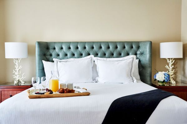 A neatly made bed with white pillows and a tray of breakfast items, flanked by lamps and flowers on bedside tables.