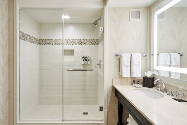 The image shows a modern bathroom with a glass shower enclosure, elegant sink, illuminated mirror, and neatly arranged towels on racks.