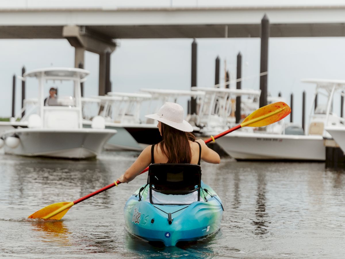 A person in a blue kayak paddles in the water near a marina with boats and a bridge visible in the background.