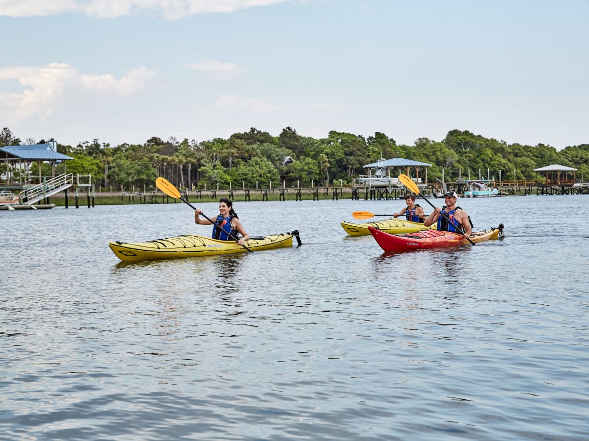 Three people kayaking on a calm body of water with trees and docks in the background.