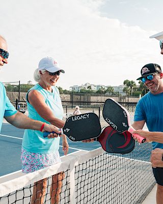Four people on a pickleball court, smiling and tapping their paddles over the net, possibly celebrating or starting a game.