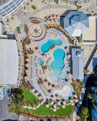 An aerial view of a resort features multiple swimming pools, lounge chairs, umbrellas, landscaped areas, and surrounding buildings.