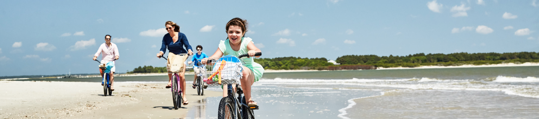 A group of people are riding bicycles on a beach with blue skies and ocean waves in the background.