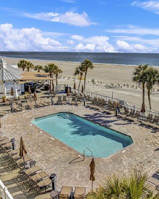 A beachside pool with surrounding lounge chairs and palm trees, next to a sandy beach and ocean under a blue sky with clouds.