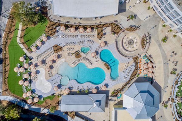 An aerial view of a resort pool area with umbrellas, lounge chairs, and surrounding buildings.