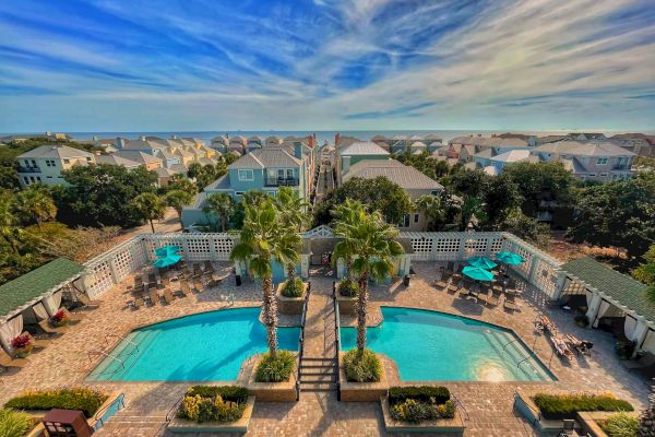A rooftop view of a swimming pool in a residential area with palm trees, lounge chairs, and blue umbrellas. Houses and greenery surround it.