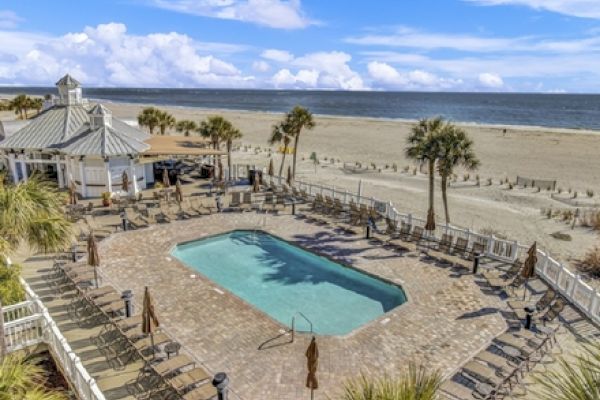 This image shows a beachside pool area with lounge chairs, palm trees, and a view of the ocean under a partly cloudy sky.