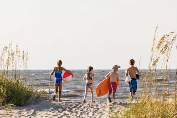 A group of people with beach gear walk toward the ocean, surrounded by sand dunes and tall grass under a clear sky.