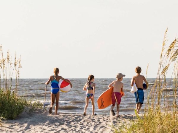 A group of people with beach gear walk toward the ocean, surrounded by sand dunes and tall grass under a clear sky.
