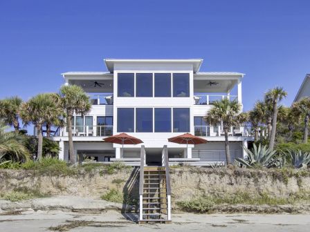 A modern beachfront house with large windows, surrounded by palm trees and plants, features a stairway leading down to the beach.