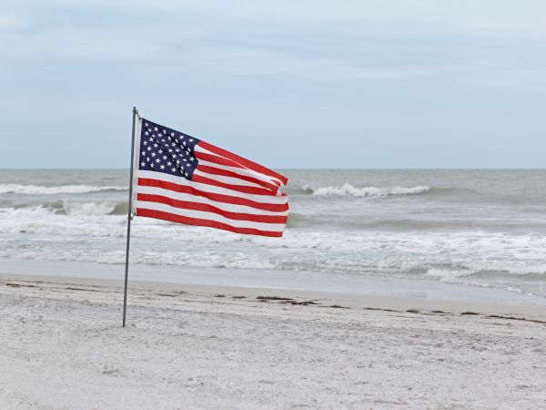 An American flag waving on a pole stands on a sandy beach with the ocean and waves in the background under a cloudy sky.