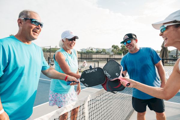 Four people smiling and touching pickleball paddles over a net, outdoors on a sunny day.