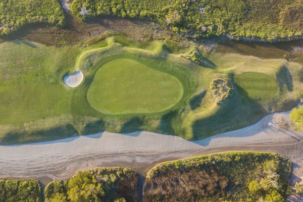 Aerial view of a golf course section with a green, sand bunker, pathways, and surrounding vegetation.