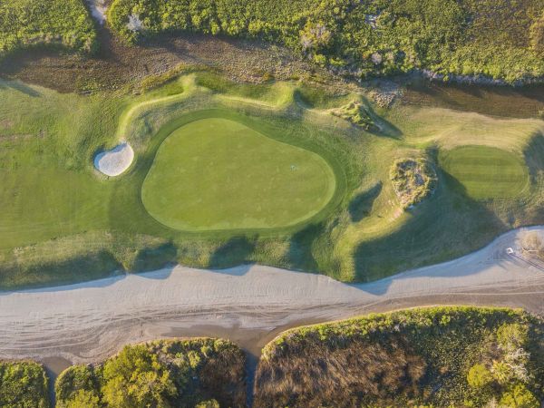 Aerial view of a golf course section with a green, sand bunker, pathways, and surrounding vegetation.