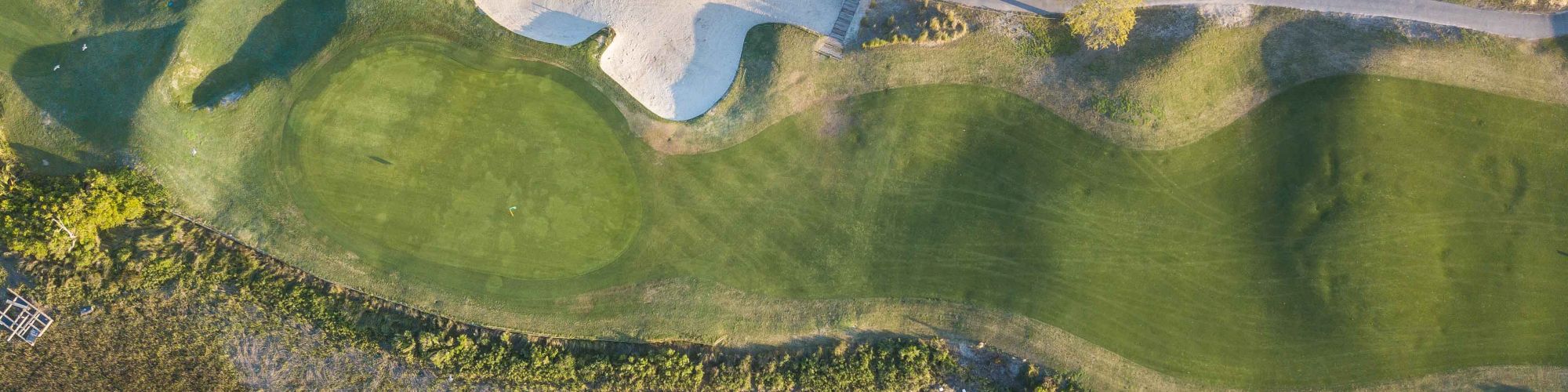 Aerial view of a golf course with green fairways, a sand bunker, and nearby trees and residences.