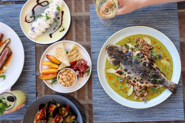 A table with a whole fish dish, assorted appetizers, desserts, and a hand holding a glass. Plates are artistically arranged.