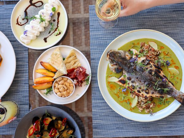 A table with a whole fish dish, assorted appetizers, desserts, and a hand holding a glass. Plates are artistically arranged.