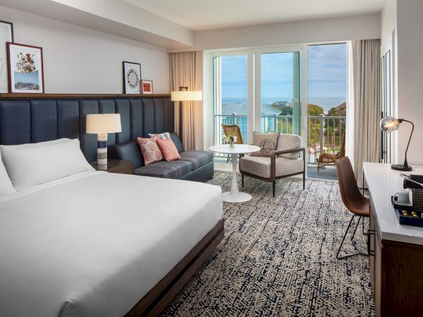 A modern hotel room with a bed, sofa, chairs, desk, and a balcony with an ocean view. Artwork decorates the walls.