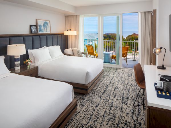 A modern hotel room with two beds, lamp, desk, and TV, features a balcony with a view of the sea and outdoor seating.