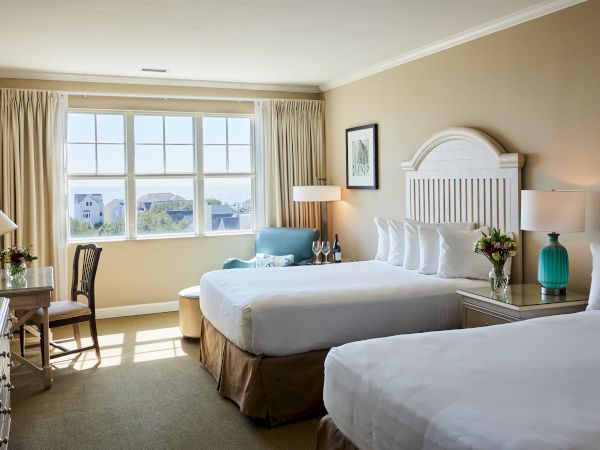 A hotel room with two beds, large window, chair, lamps, wine, and coastal view. Decor includes flowers and framed art on the wall.