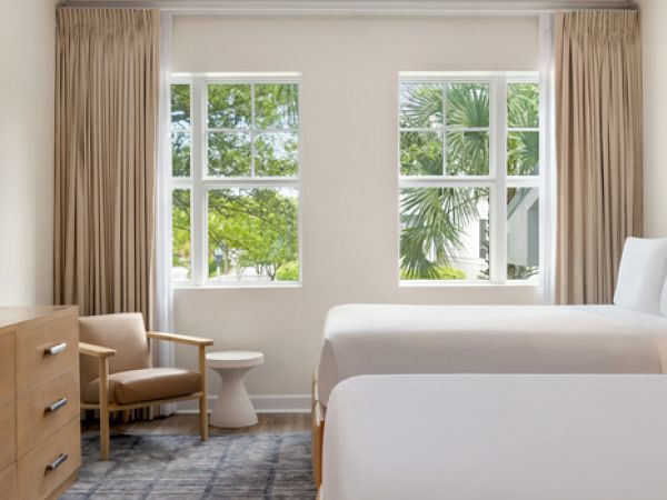 The image shows a hotel room with two beds, a TV, a wooden dresser, a chair, and large windows with a view of greenery outside.