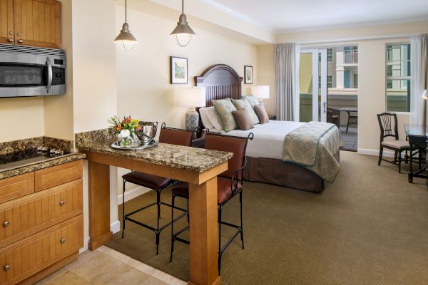 The image shows a cozy hotel room with a bed, kitchenette, chairs, and a small table, featuring a warm and inviting ambiance.