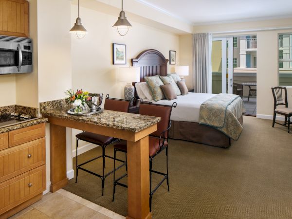 The image shows a cozy hotel room with a bed, kitchenette, chairs, and a small table, featuring a warm and inviting ambiance.