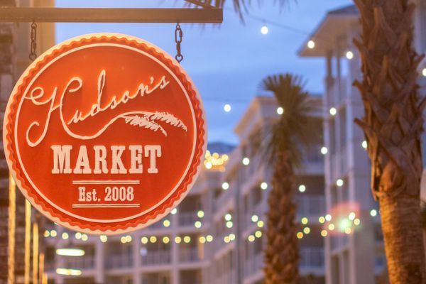An outdoor view of a market sign reading “Hudson's MARKET Est. 2008” with strings of lights and palm trees in the background, evening setting.