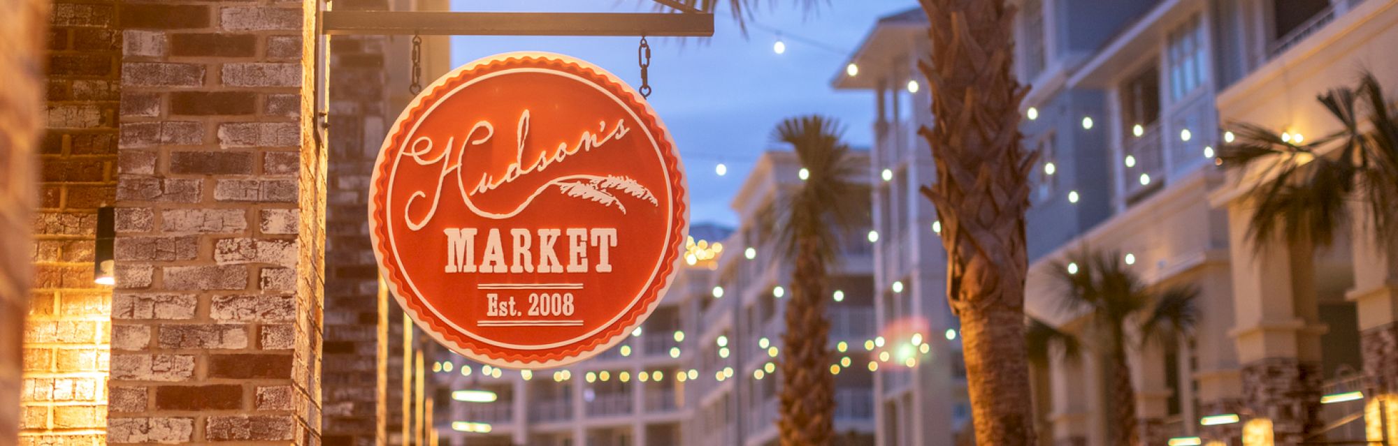 The image shows a sign for "Hudson's Market" with string lights and palm trees in the background.