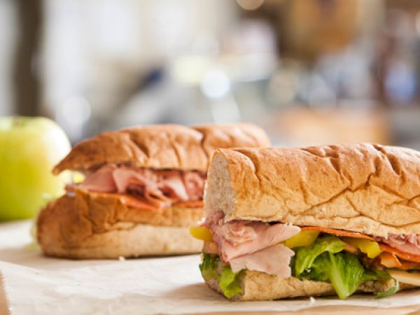 Two sub sandwiches with lettuce, ham, cheese, and other fillings are placed on paper. An apple is blurred in the background.