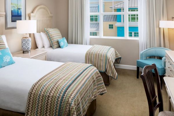 The image shows a hotel room with two beds, patterned bedding, a desk, chairs, lamps, and a window with a view of colorful buildings.
