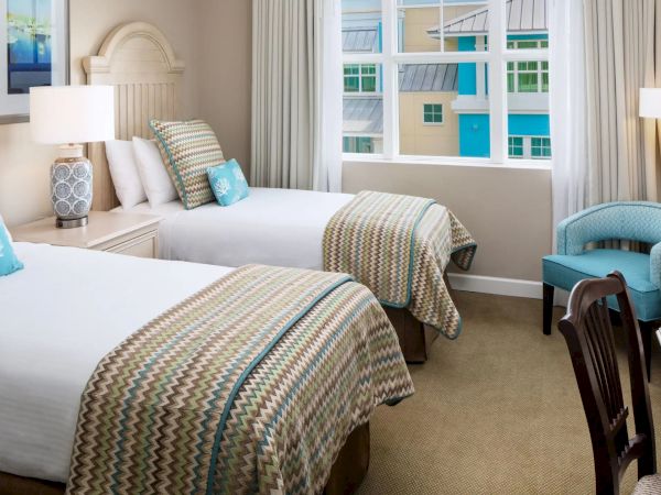 The image shows a hotel room with two beds, patterned bedding, a desk, chairs, lamps, and a window with a view of colorful buildings.