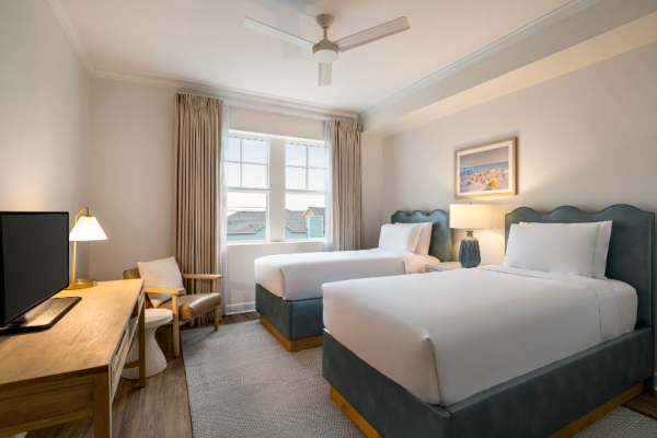 The image shows a hotel room with two beds, a desk, a chair, and a TV. There are curtains and a ceiling fan.