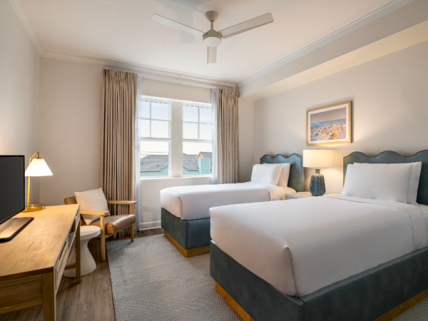 The image shows a hotel room with two beds, a desk, a chair, and a TV. There are curtains and a ceiling fan.