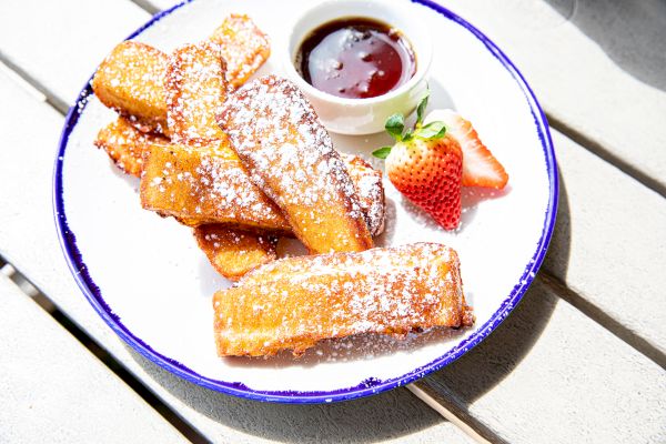 The image shows a plate of French toast sticks dusted with powdered sugar, accompanied by a small cup of syrup and a sliced strawberry.