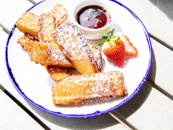 The image shows a plate of French toast sticks dusted with powdered sugar, accompanied by a small cup of syrup and a sliced strawberry.