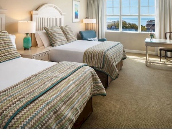 The image shows a hotel room with two beds, a desk, a chair, lamps, and a window with a view, designed in a cozy, neutral style.