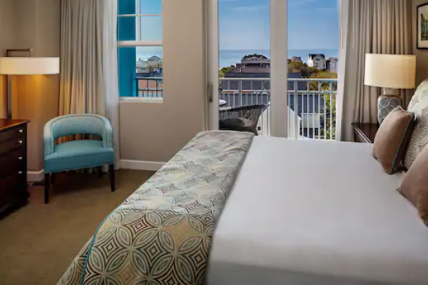 A cozy hotel room features a bed, TV, and armchair. Large windows offer a view of buildings and the sea beyond the balcony.