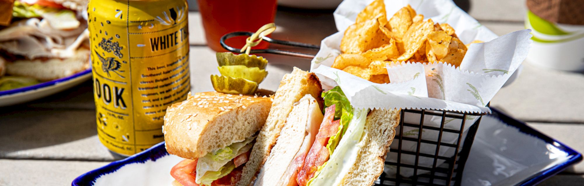 A sandwich with lettuce, tomato, and turkey, served with a basket of chips and pickles, alongside a canned drink on a tray.