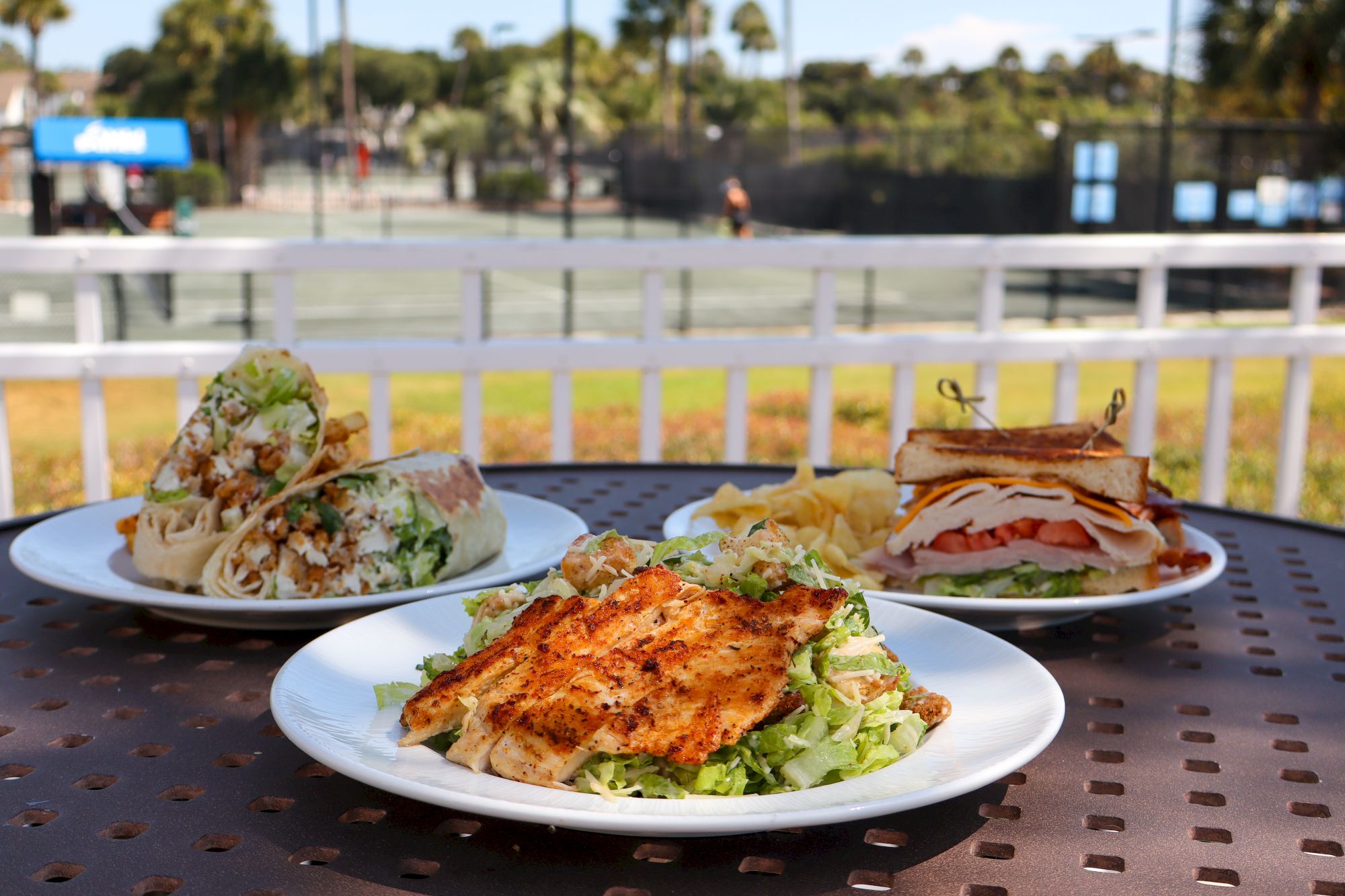 The image shows plates of food including salads and sandwiches on a table outdoors, with a tennis court visible in the background.