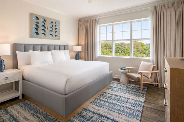 A well-decorated bedroom with a bed, lamps, artwork, and a window with a scenic view is shown.