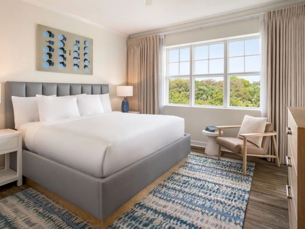 A well-decorated bedroom with a bed, lamps, artwork, and a window with a scenic view is shown.