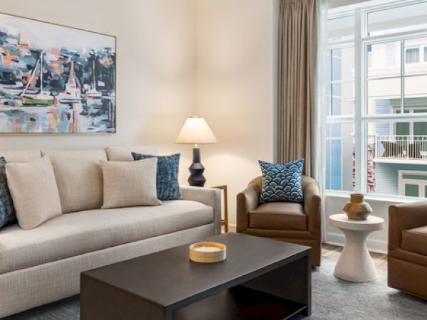 A modern living room with a sofa, two armchairs, a coffee table, lamps, abstract artwork, and large windows with a view.