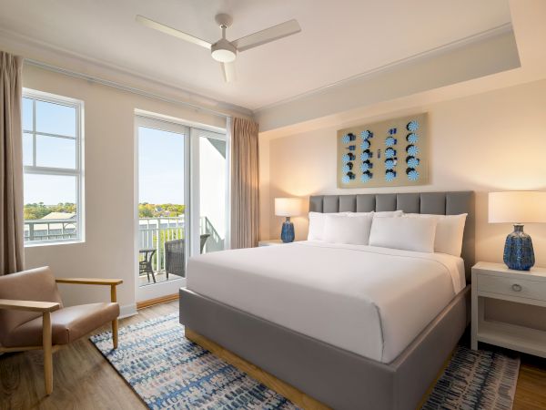 A modern bedroom with a grey bed, white bedding, blue lamps, art on the wall, and a balcony view.