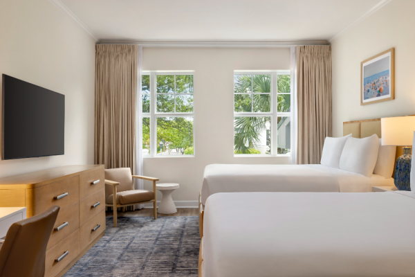 A hotel room with two beds, a TV, dresser, chair, lamps, curtains, and artwork. Windows show trees outside, with a neutral color scheme.