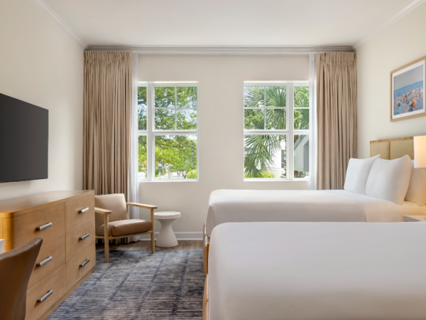 A hotel room with two beds, a TV, dresser, chair, lamps, curtains, and artwork. Windows show trees outside, with a neutral color scheme.