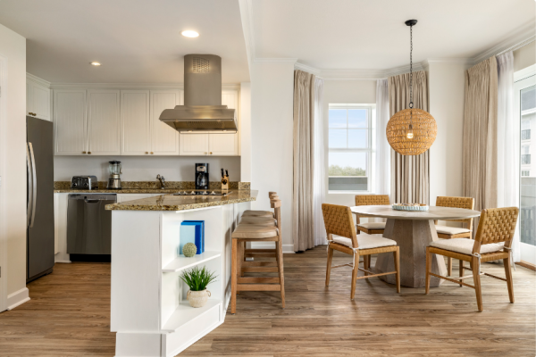 The image shows a modern kitchen and dining area with wooden floors, granite countertops, a round table, and natural lighting.