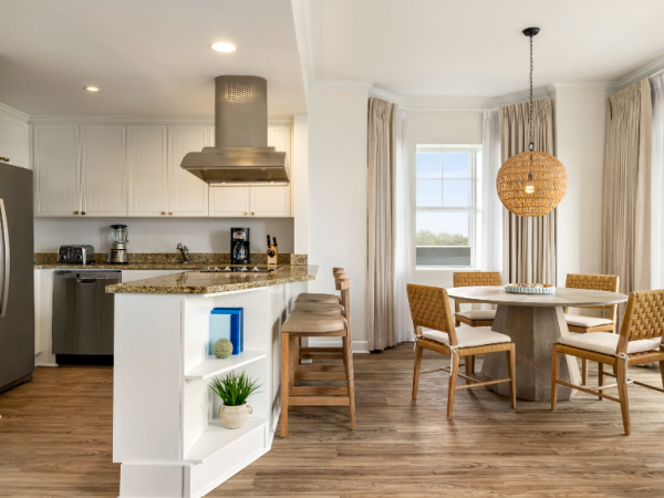 The image shows a modern kitchen and dining area with wooden floors, granite countertops, a round table, and natural lighting.